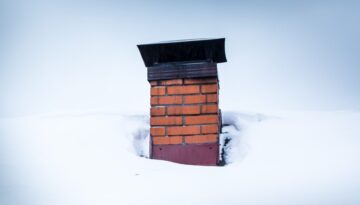 A snow-covered roof with a brick chimney and black chimney cap.