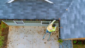 Man in a reflective vest standing on a ladder and performing gutter maintenance on a house.