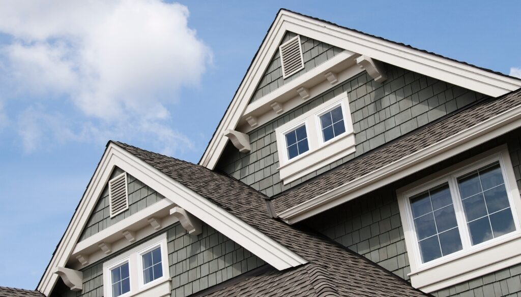 The top half of a grey house with white trim and asphalt shingles under a blue sky.