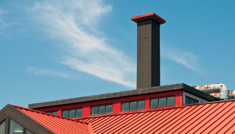 A commercial building with a red roof and black commercial chimney with a blue sky behind it.