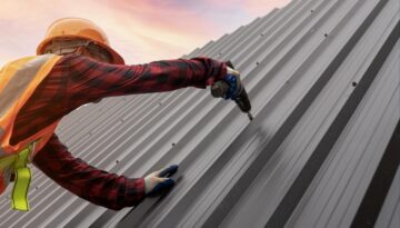 Worker in safety gear installing a sleek metal roof under a vibrant sunset sky.