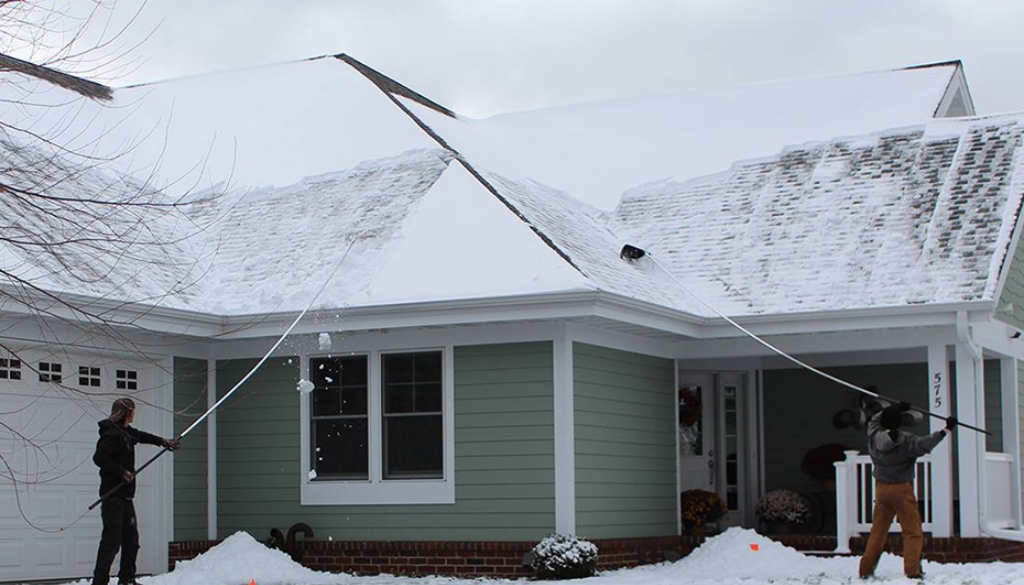 Two people using roof rakes to safely remove snow from a residential roof during winter