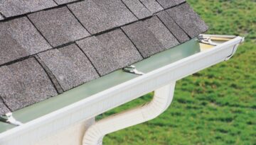 A close-up of a home’s gutter and downspout system installed below an asphalt shingle roof to direct rainwater away from the foundation.