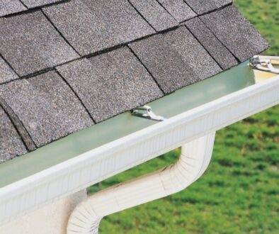 A close-up of a home’s gutter and downspout system installed below an asphalt shingle roof to direct rainwater away from the foundation.