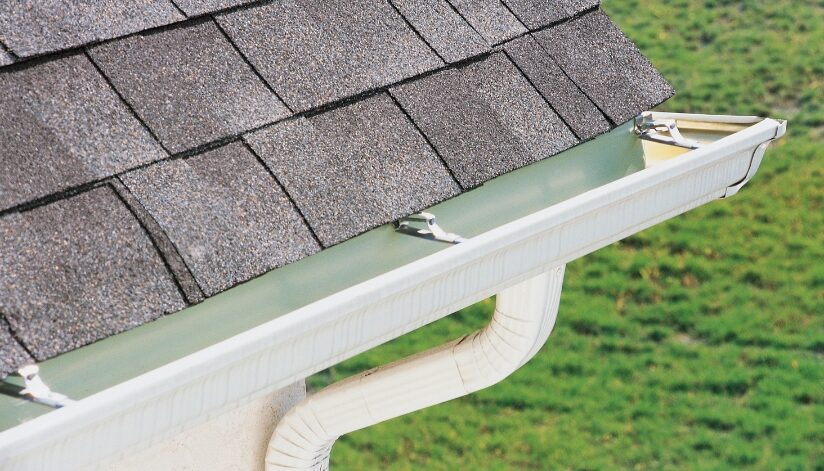 A close-up of a home’s gutter and downspout system installed below an asphalt shingle roof to direct rainwater away from the foundation.