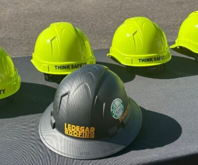 A row of bright yellow hard hats labeled “Think Safety” next to a black Ledegar Roofing hard hat with an OSHA-trained roofing safety sticker.