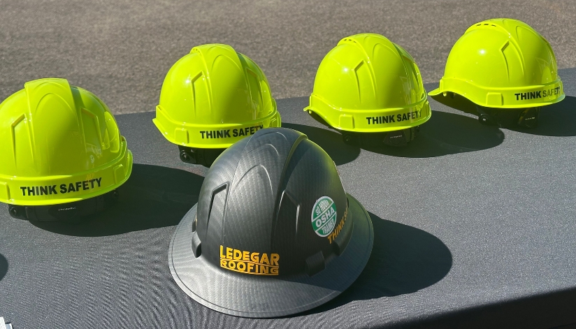 A row of bright yellow hard hats labeled “Think Safety” next to a black Ledegar Roofing hard hat with an OSHA-trained roofing safety sticker.